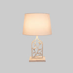 A white metal table lamp with a bird and leaf design on the base and a round shade.